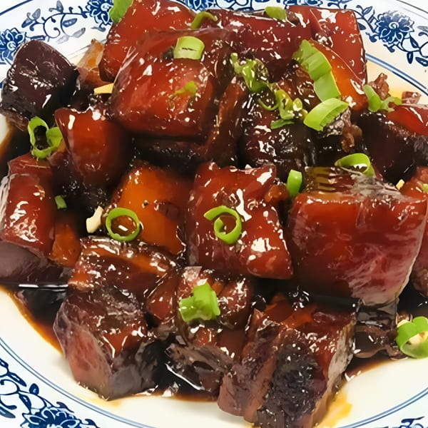 Braised pork belly, glistening with a dark sauce and garnished with chopped green onions, in a blue and white patterned bowl.